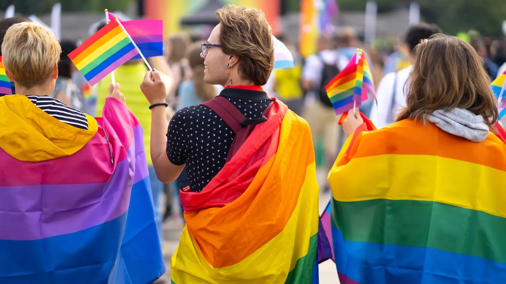 Wrapped in bisexual flag and pride flags this trio are waving small pride flags and watching a gay pride event