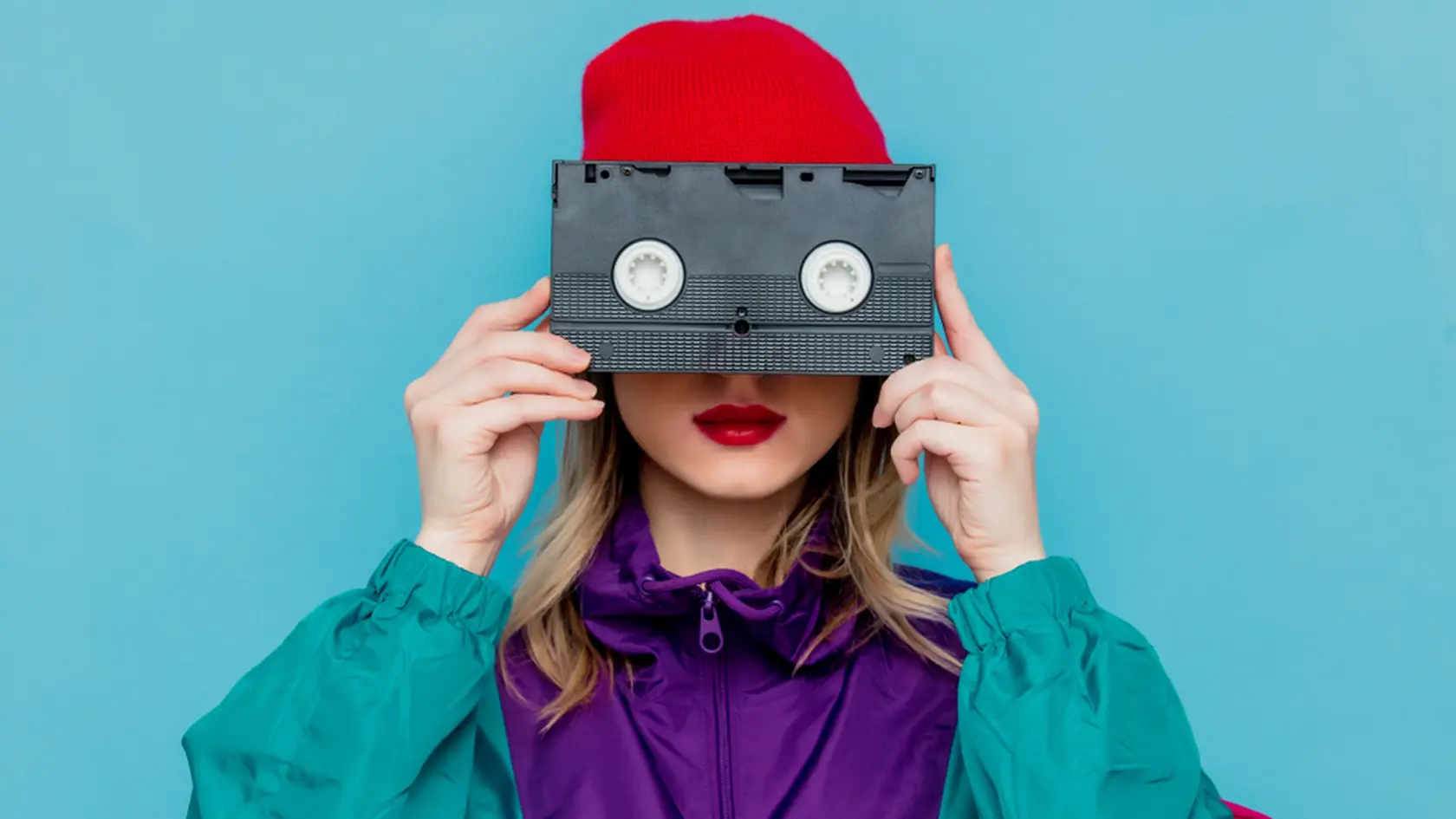 Portrait of a woman in red hat, sunglasses and suit of 90s with VHS cassette on blue background.
