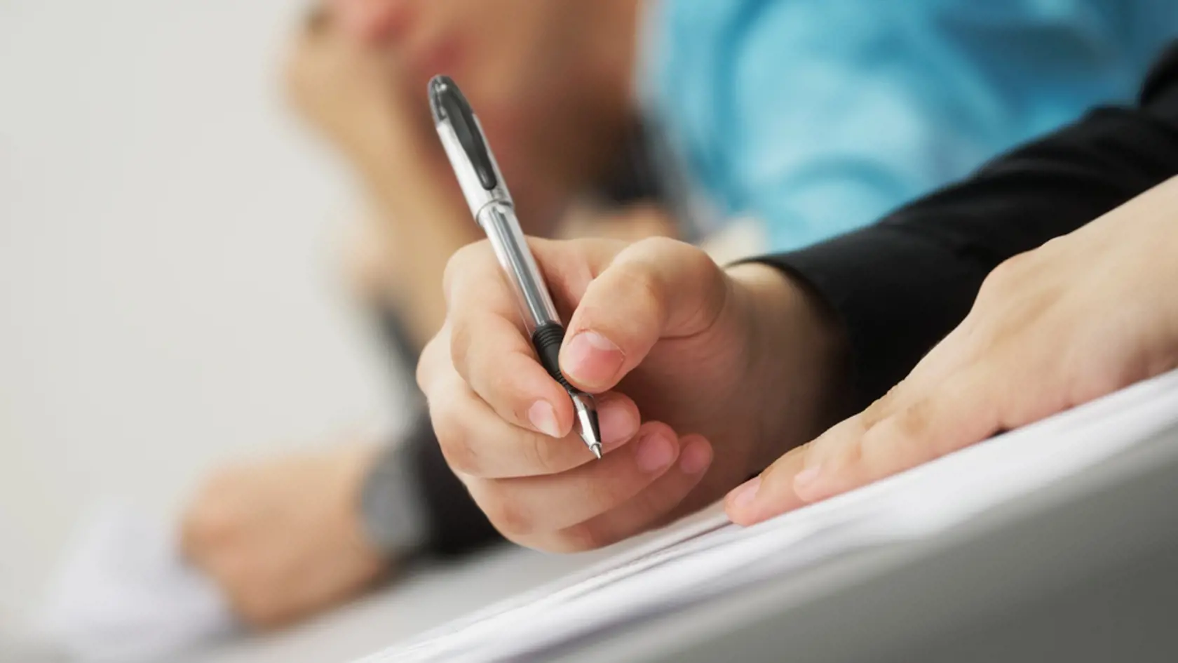 A young man, a student or schoolchild, writes a dictation, an essay or solves an educational problem, sitting at a desk next to other students. Photo. Selective focus. No face. Close-up