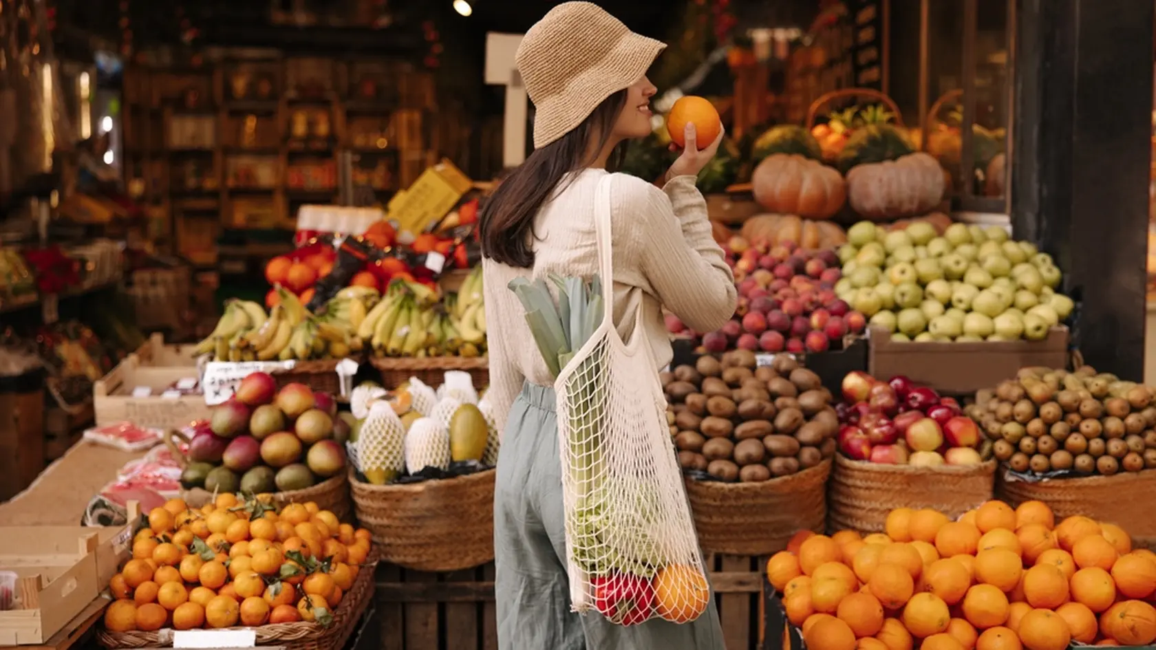 Fair-skinned young girl holds orange standing with her back to camera in fruit street shop. Brunette wears hat, blouse, pants and string bag. Concept shopping