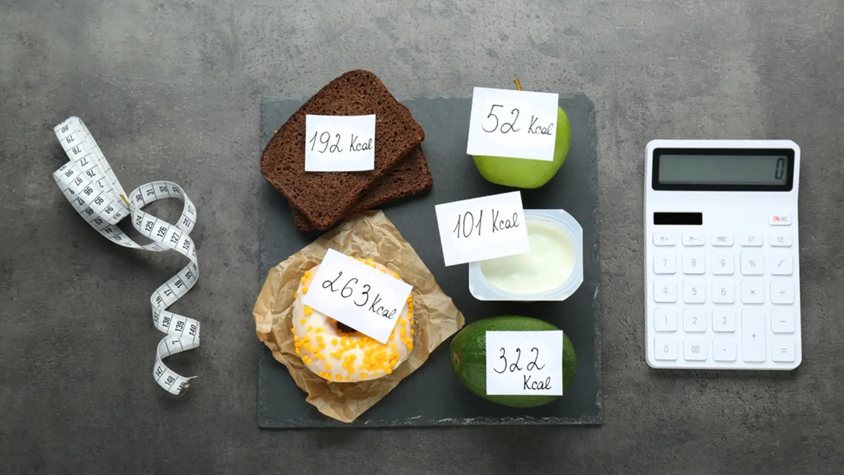 Calculator and food products with calorific value tags on dark grey table, flat lay. Weight loss concept