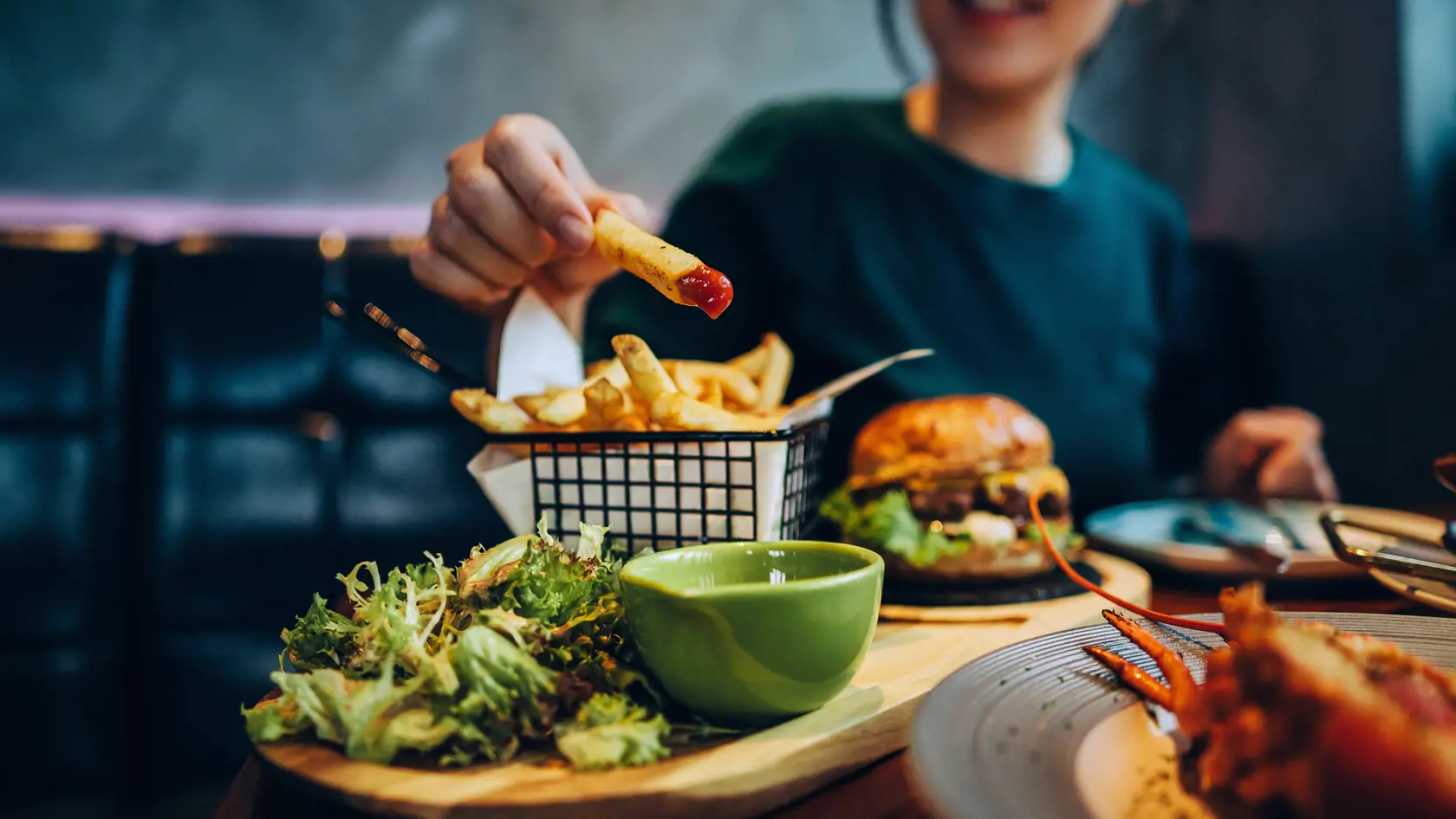 Cropped shot of young Asian woman eating freshly made delicious burger, dipping fries in ketchup on the dining table. Enjoying a feast in the restaurant. People, food and lifestyle