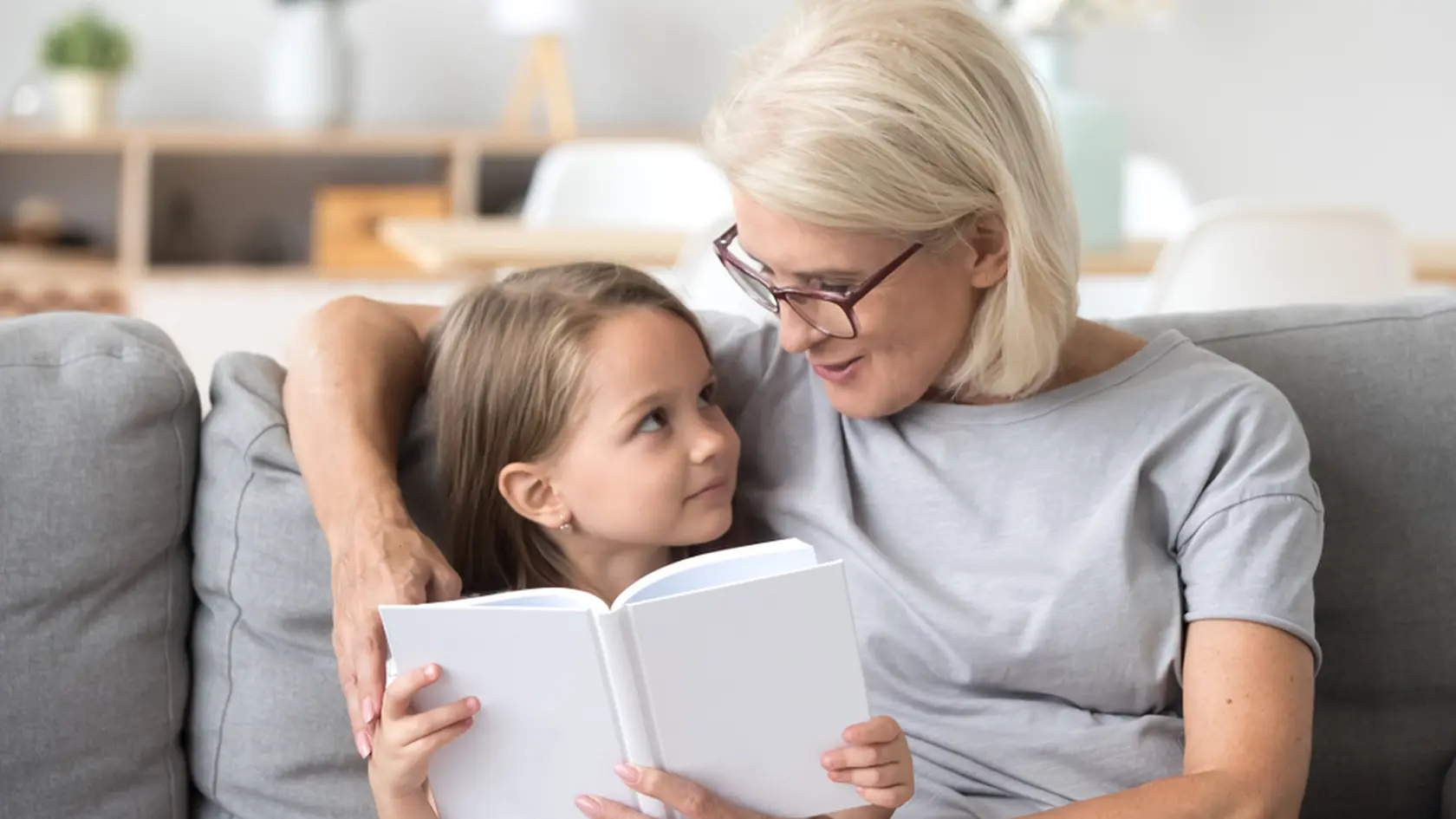 Loving grandmother teaching granddaughter holding book sitting on sofa, grandma baby sitter embracing kid girl reading fairytale to cute child, nanny granny telling story to preschool grandchild