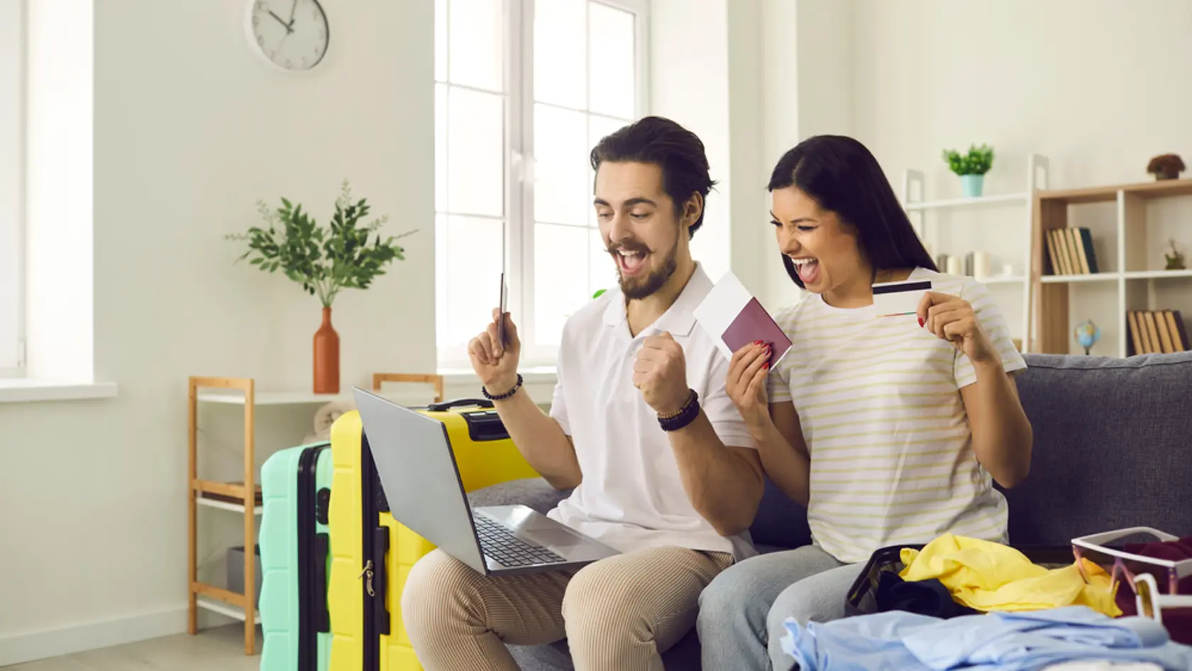 Happy couple getting ready for tour vacation trip pay for cheap hotel online. Excited boyfriend and girlfriend sitting on sofa with laptop, credit card, passports, flight tickets and travel suitcase
