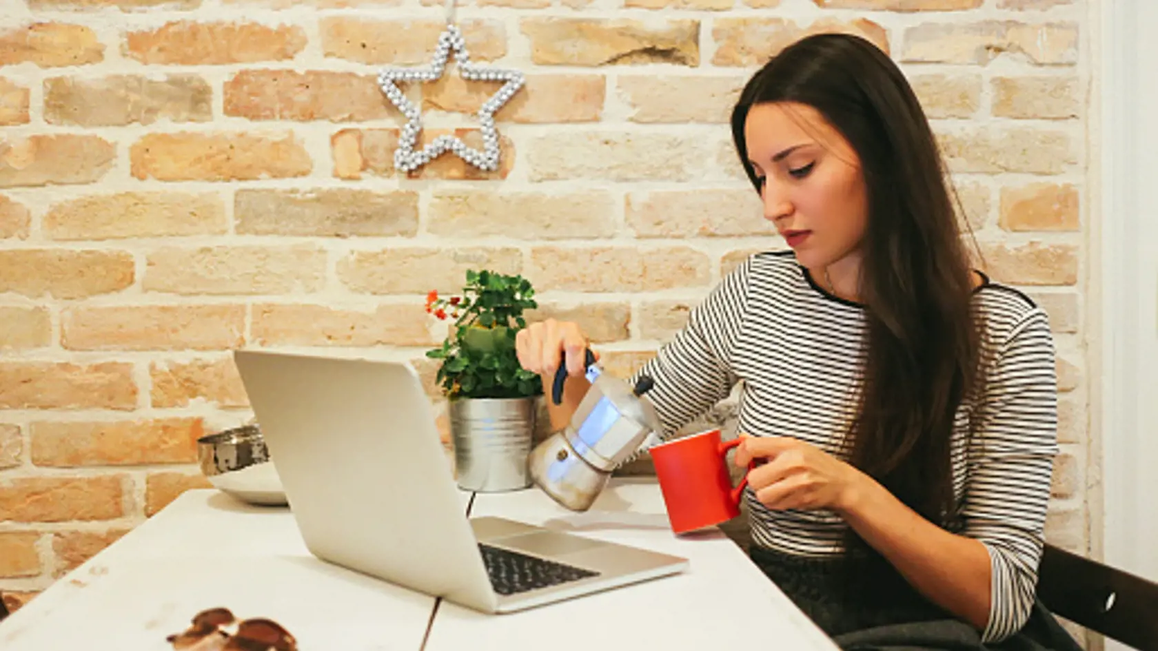 Vintage warm toned image of a young female novelist or a blogger, typing on the laptop keyboard in her bright apartment or an office space, sitting on the chair behind the desk. She's having a cup of hot coffee or tea while she's working or just browsing the web. Lifestyle - freelancer work concept in a casual environment.