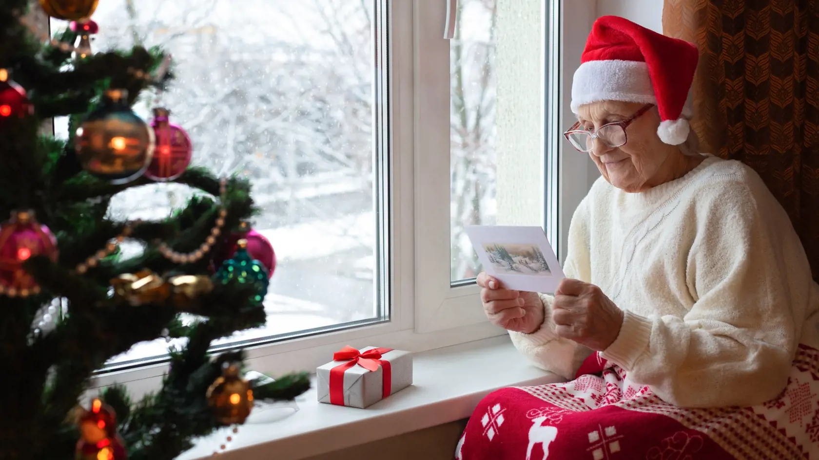 Elderly Caucasian woman Santa hat sits window with glasses her face and reads greeting card.Christmas eve, gift, Christmas tree.Concept: Christmas holidays, New Year, caring for elderly, the lonely