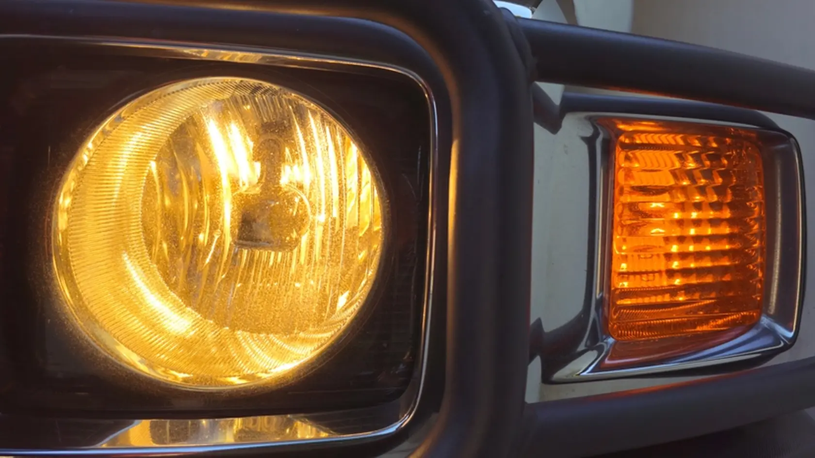 A close-up of a classic off-road vehicle's glowing headlight and turn signal, featuring a rugged metal grille. The warm amber and yellow lights create a nostalgic, adventure-ready atmosphere.