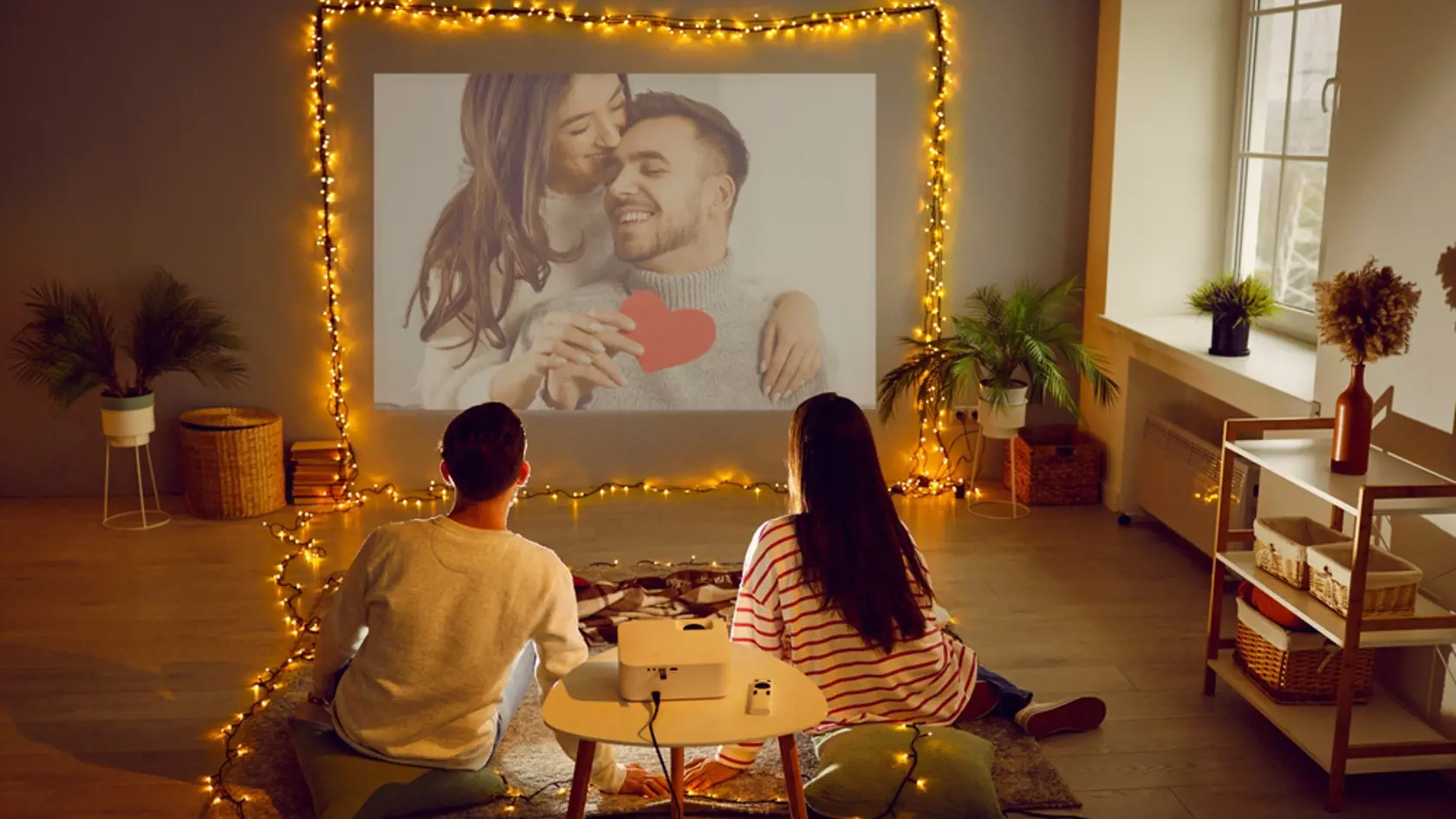 Romantic couple having Valentine's Day date at home, using modern projector in living room interior with beautiful fairy lights, watching movie, looking at digital photos, enjoying memories. Back view
