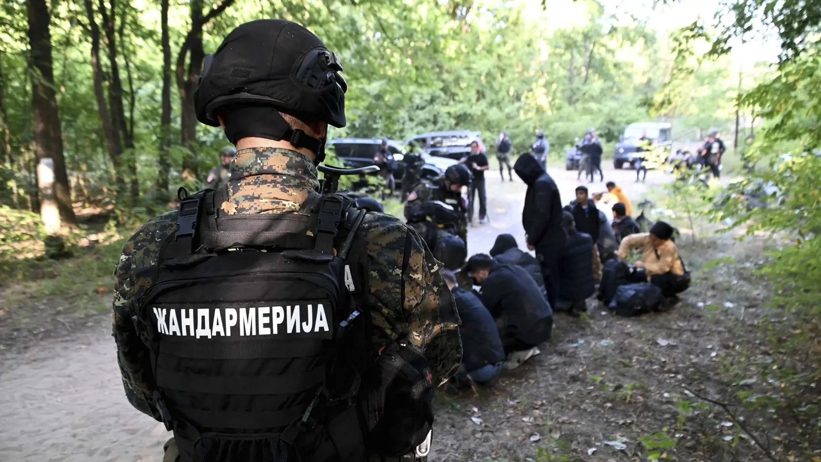 Srbský policajt stráži zadržaných migrantov, ktorí sedia na zemi na neznámom mieste v blízkosti hraníc s Maďarskom v utorok 12. septembra 2023.