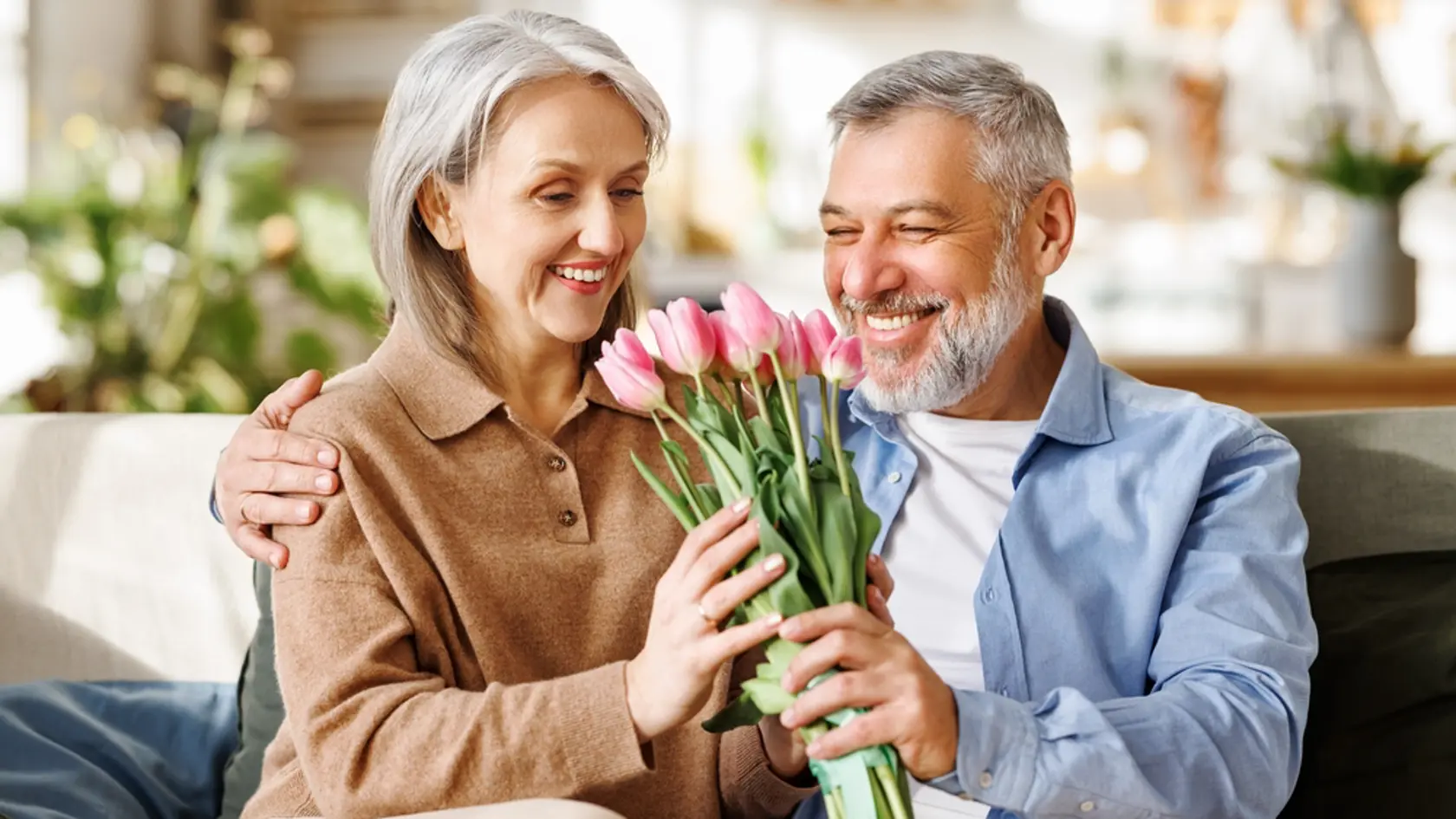 Happy senior woman getting romantic present from husband on Valentines Day while sitting on sofa at home, selective focus. Pleased elderly female receiving bouquet of spring flowers on March 8