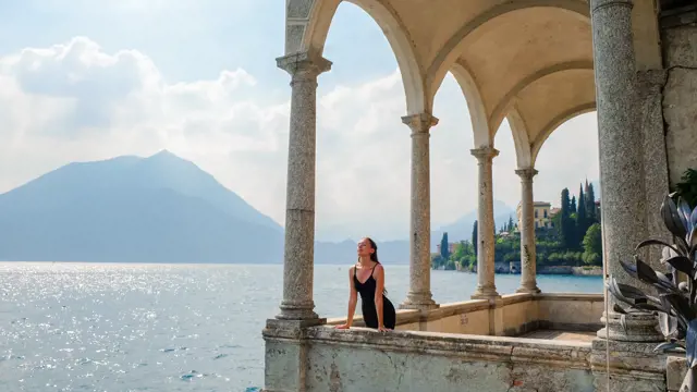 a young woman in a black dress standing on the old balcony of villa Monstera looking at the lake Como Bellagio amazing view. Italian Alps summer vacation rental. Aged beautiful architecture booking.