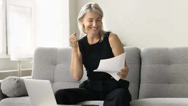 Happy successful mature investor woman celebrating profit, income from investment, retirement savings, reading financial paper document, making winner hand, smiling, sitting at laptop at home